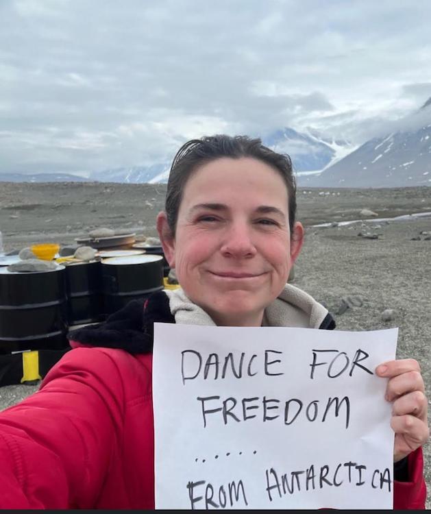 head shot of a smiling woman in Antartica, behind her are beautiful clouds, a range of mountains, and closer a grouping of steel drums... she holds a sign saying dance for freedom...